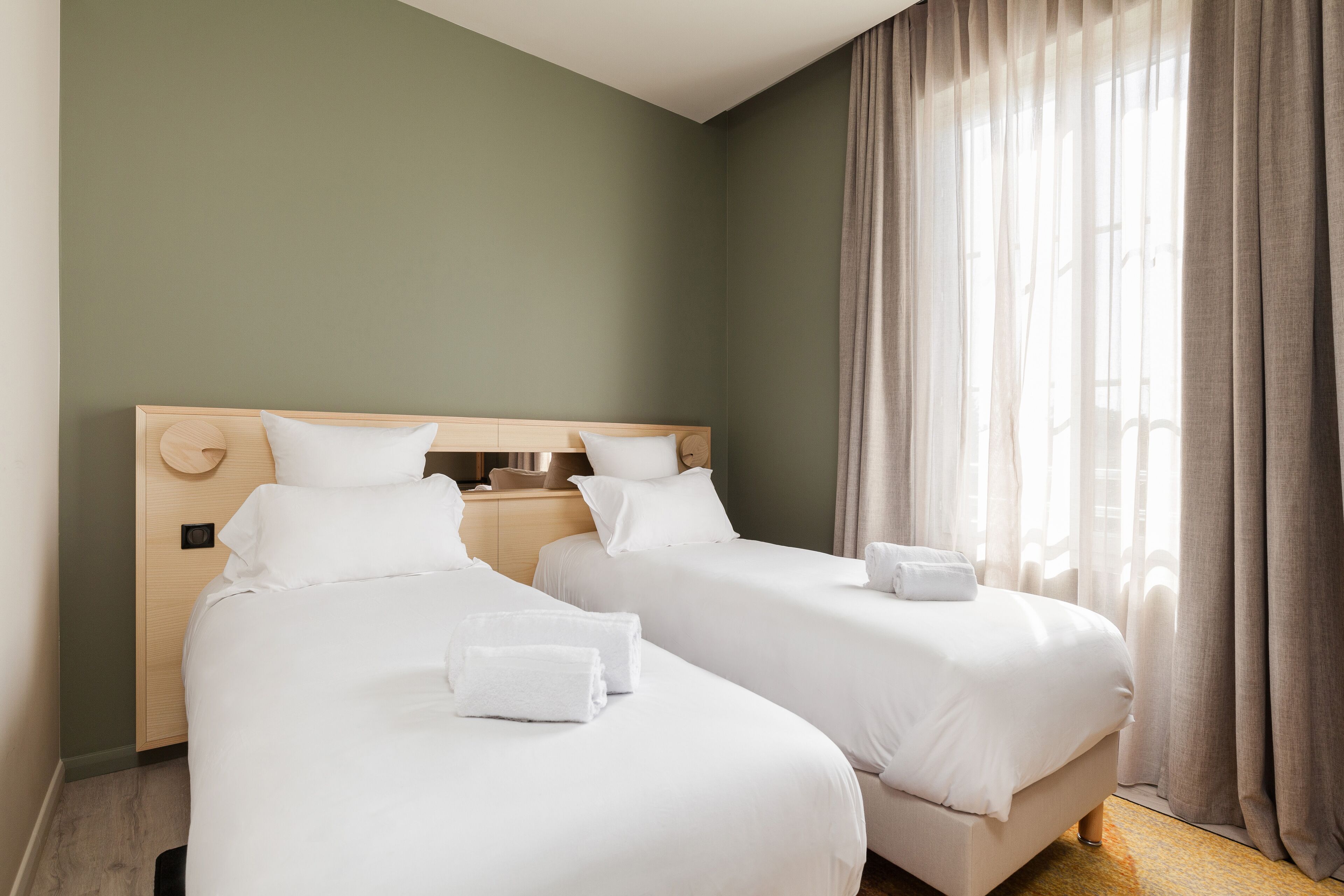 A hotel room with two single beds, a wooden headboard, white bedding, and a large window with curtains.