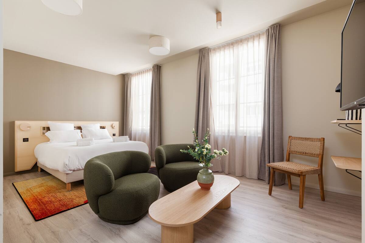 A modern hotel room with a bed, two armchairs, a wooden table, and a TV.