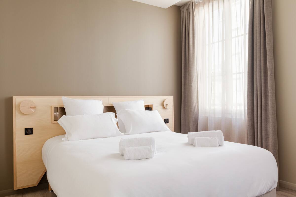 A neatly made bed with white linens and two towels, a wooden headboard, and a window with sheer curtains.