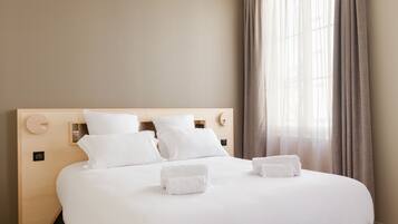 A neatly made bed with white linens and two towels, a wooden headboard, and a window with sheer curtains.