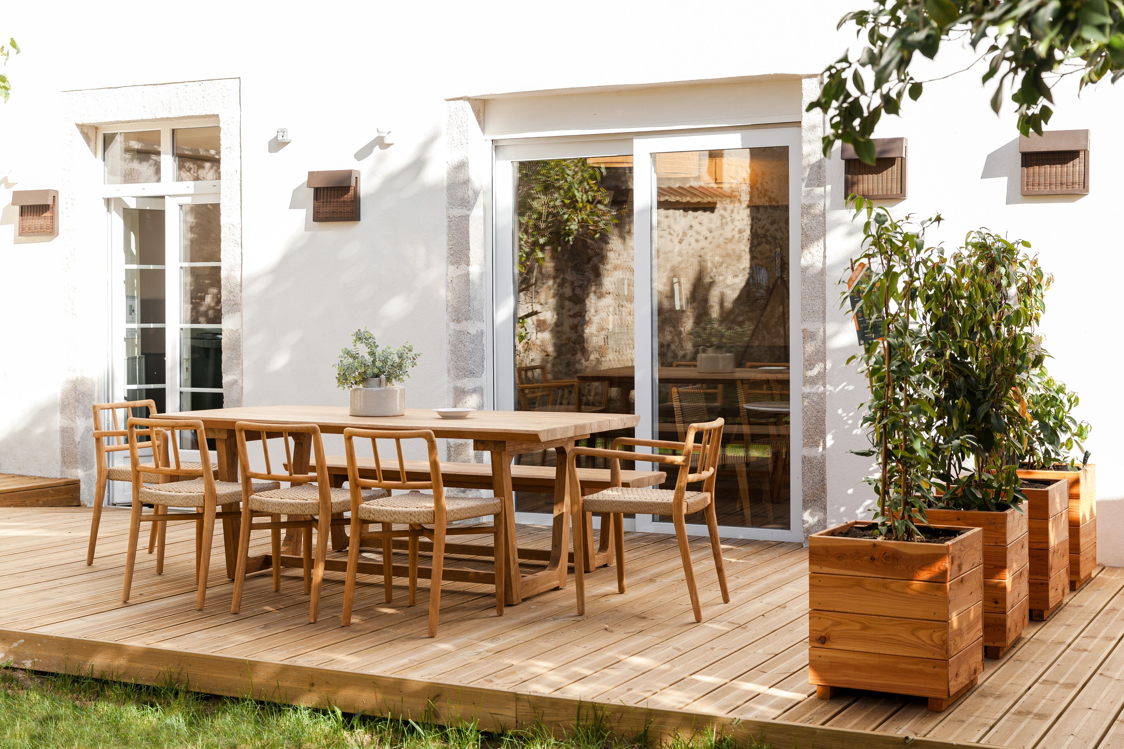 Una terraza de madera al aire libre con una mesa y sillas de comedor, rodeada de plantas en macetas.