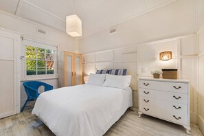 Quarto com cama, cômoda, cadeira e janela com vista para o verde.