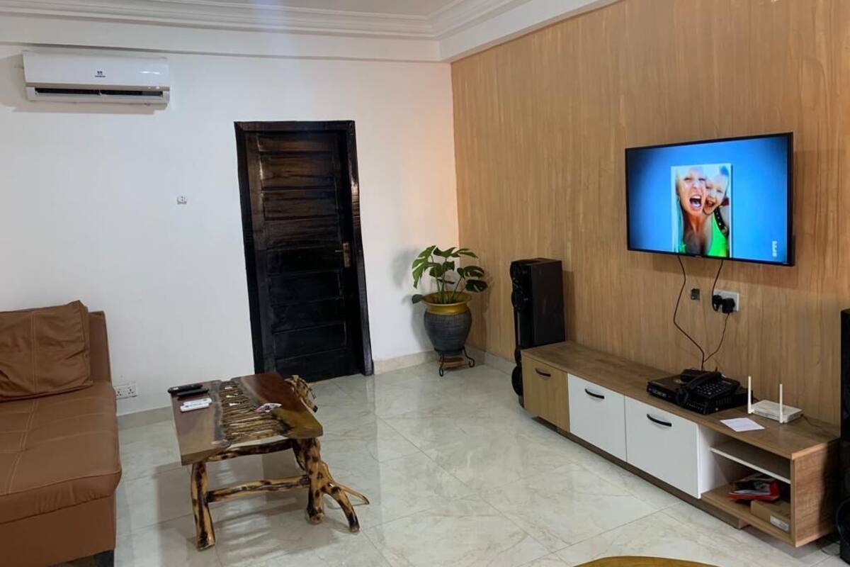 A living room with a brown sofa, a wooden coffee table, a TV mounted on a wooden wall, and a potted plant.