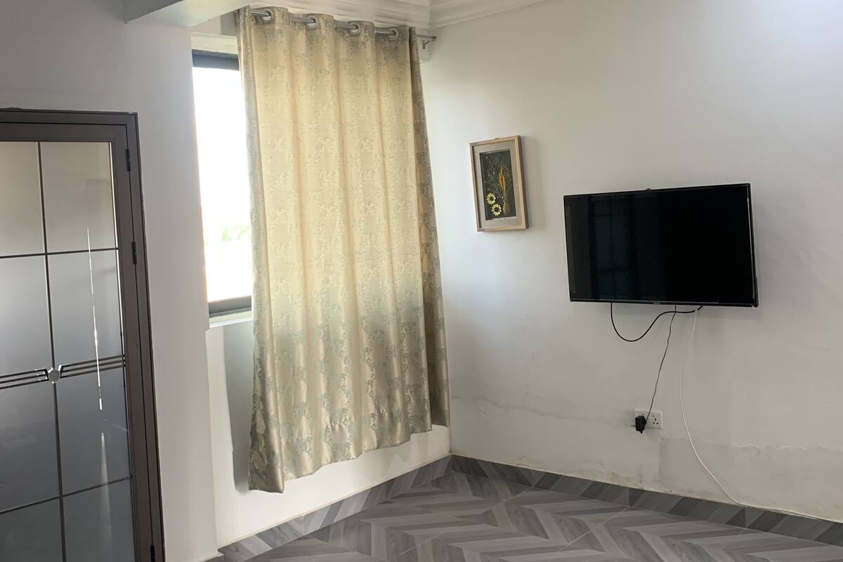 A bedroom with a bed, a TV mounted on the wall, a window with curtains, and a door.