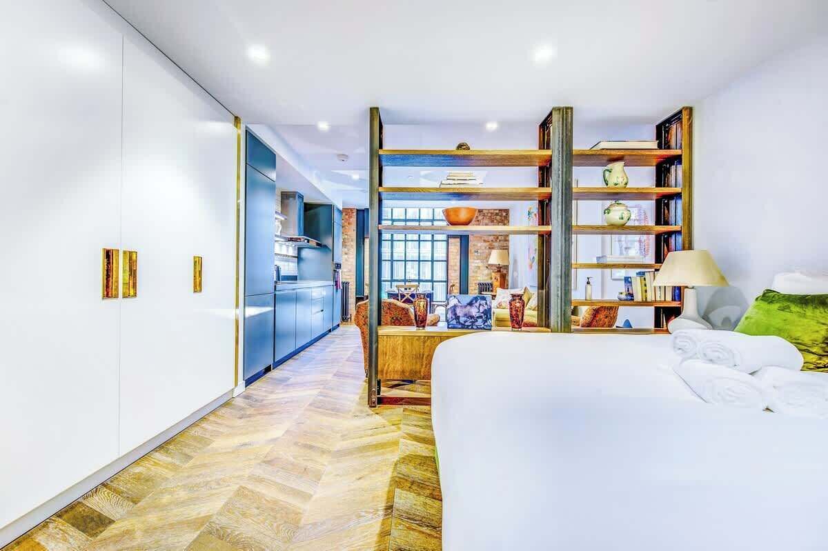 A modern hotel room with a large bed, a wooden bookshelf, a kitchen area, and a view of the city.