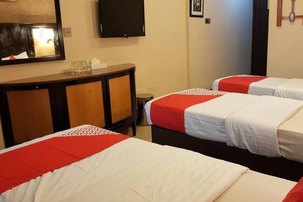A hotel room with three beds, each with red and white bedding, a wooden dresser, a flat-screen TV, and a small trash can.