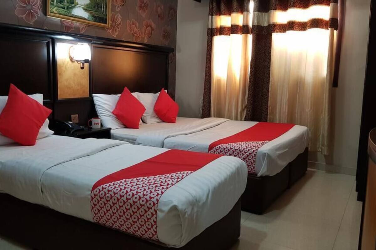 Two single beds with red pillows, a wooden headboard, a nightstand with a phone and a cup, and a window with curtains.