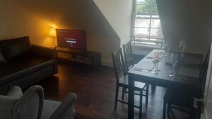 Un salon avec un canapé marron foncé, un téléviseur posé sur un meuble et un coin repas avec une table mise pour deux personnes.