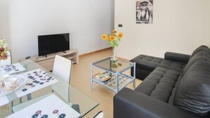 A living room with a TV, a sofa, a coffee table with a vase of flowers, and a dining area with a table set for two. - Apartamento de Vacaciones (Alacant)