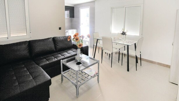 A modern living room with a black sofa, a glass coffee table, and a dining area with a white table and chairs. - Apartamento de Vacaciones (Alacant)