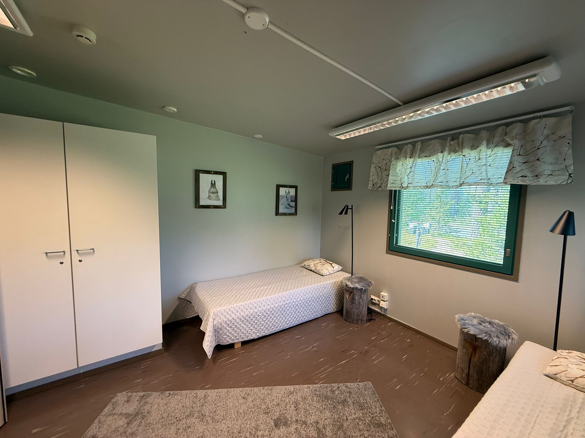A bedroom with a single bed, a wardrobe, a window with curtains, a lamp, and a small stool.