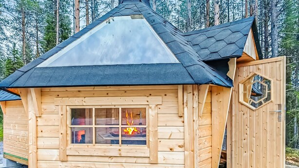 Una cabaña de madera con techo inclinado, una ventana que muestra una chimenea encendida en su interior, y una puerta con un diseño geométrico.