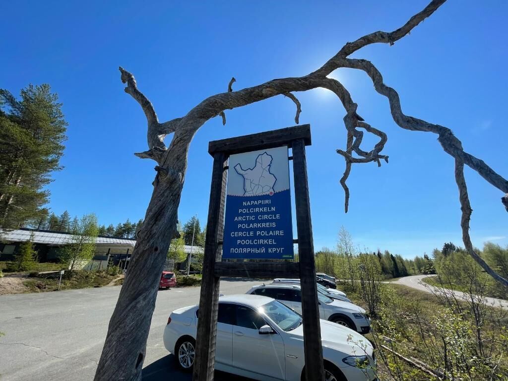 A signboard with multiple names for the Arctic Circle, a parking area with cars, and a clear blue sky.