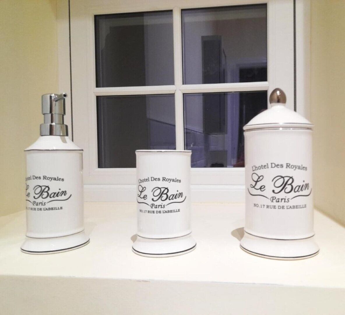 Three toiletry containers labeled 'Le Bain Paris' on a white shelf in front of a window.