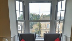Una habitación con dos sillones grises y un reposapiés a juego, una mesita con lámpara, y un ventanal con vistas a edificios y árboles.