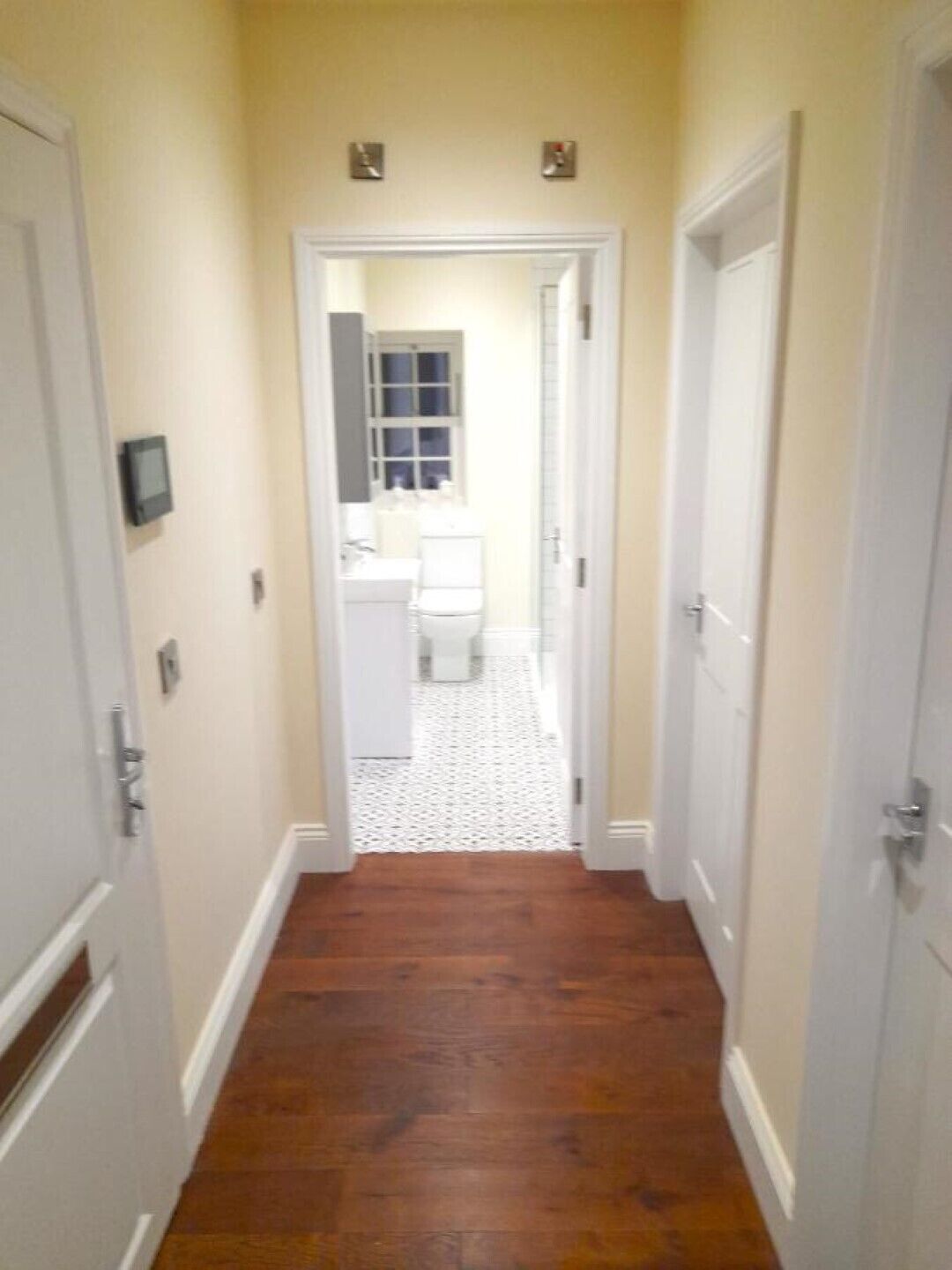 A narrow hallway with wooden flooring leading to a bathroom with a white toilet and sink.