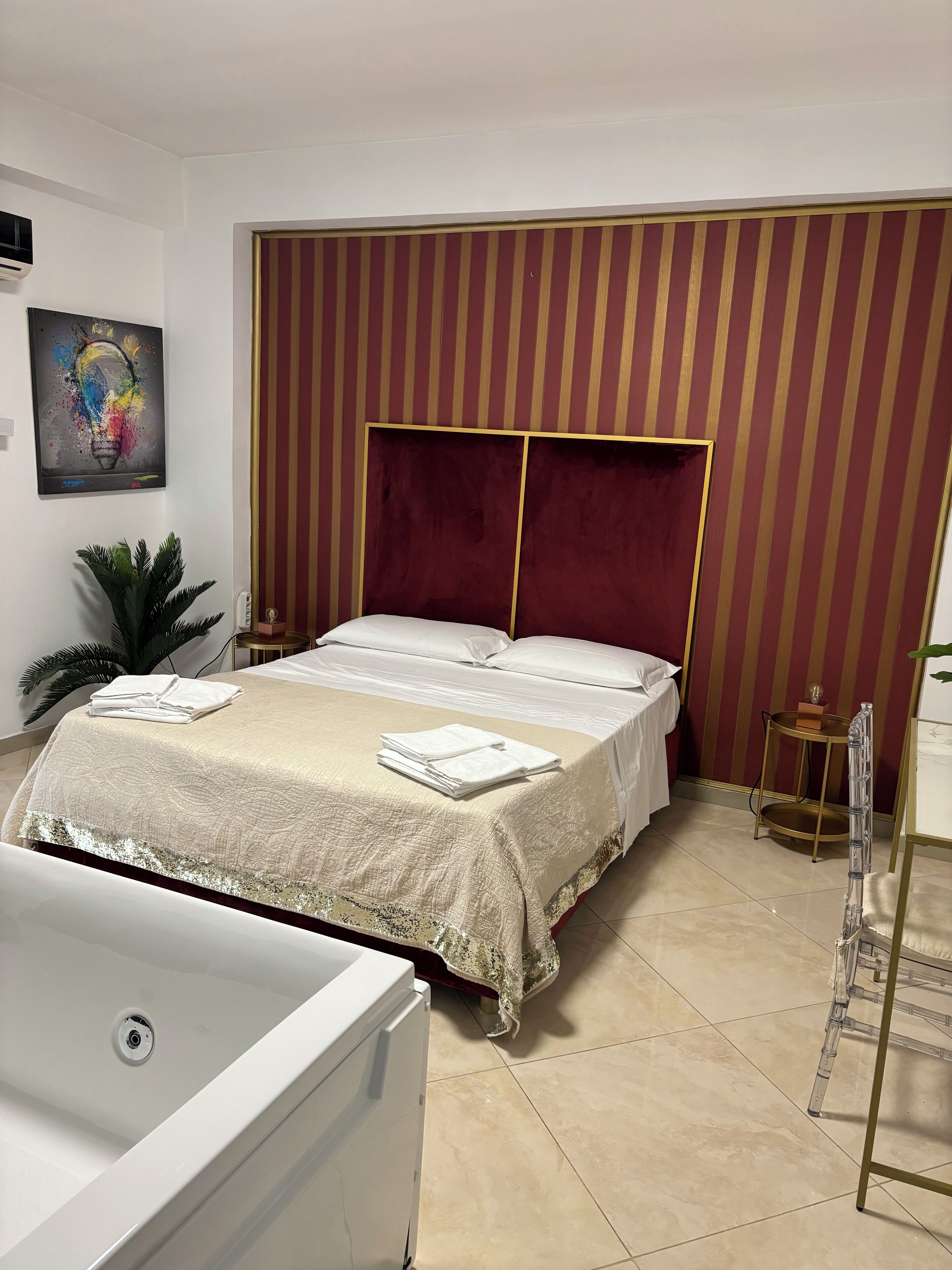 A neatly made bed with a white comforter and pillows, a large potted plant, a wall-mounted air conditioner, and a painting of a light bulb.