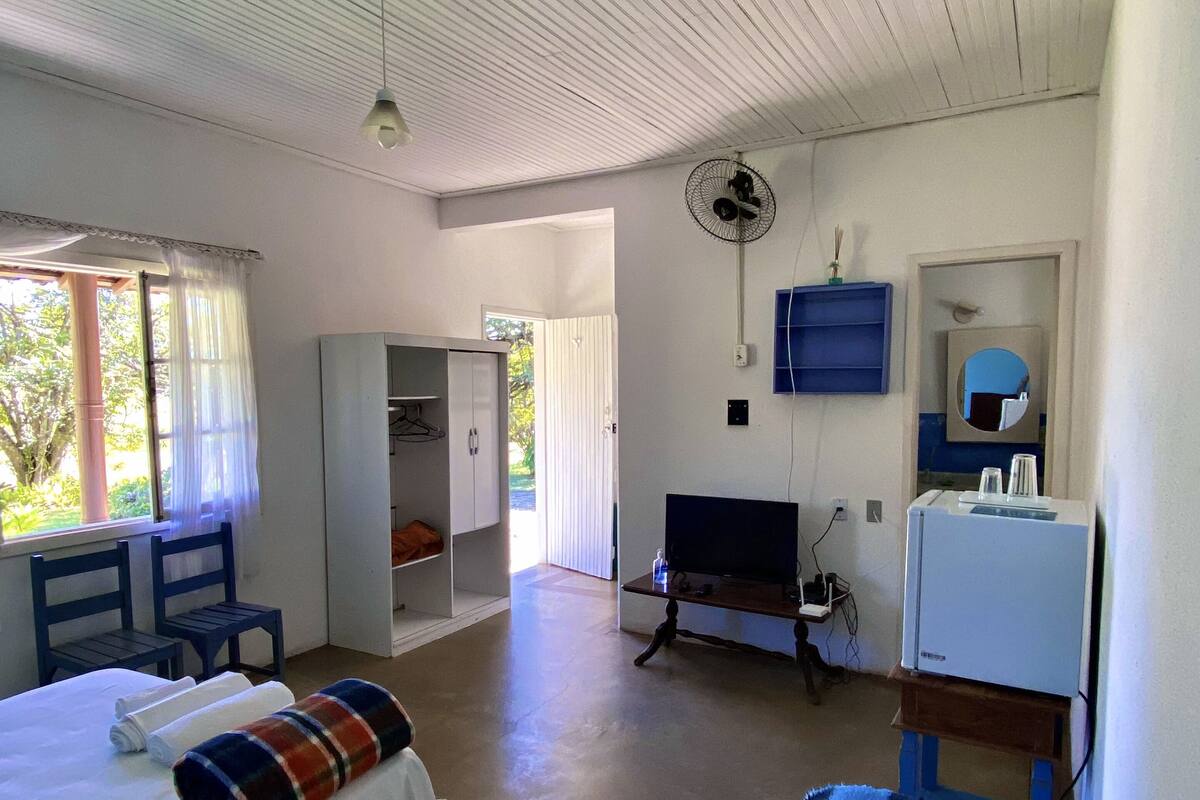 Ein Zimmer mit einem Bett, einem Sessel, einem Schreibtisch, einem Fernseher und einem Ventilator.