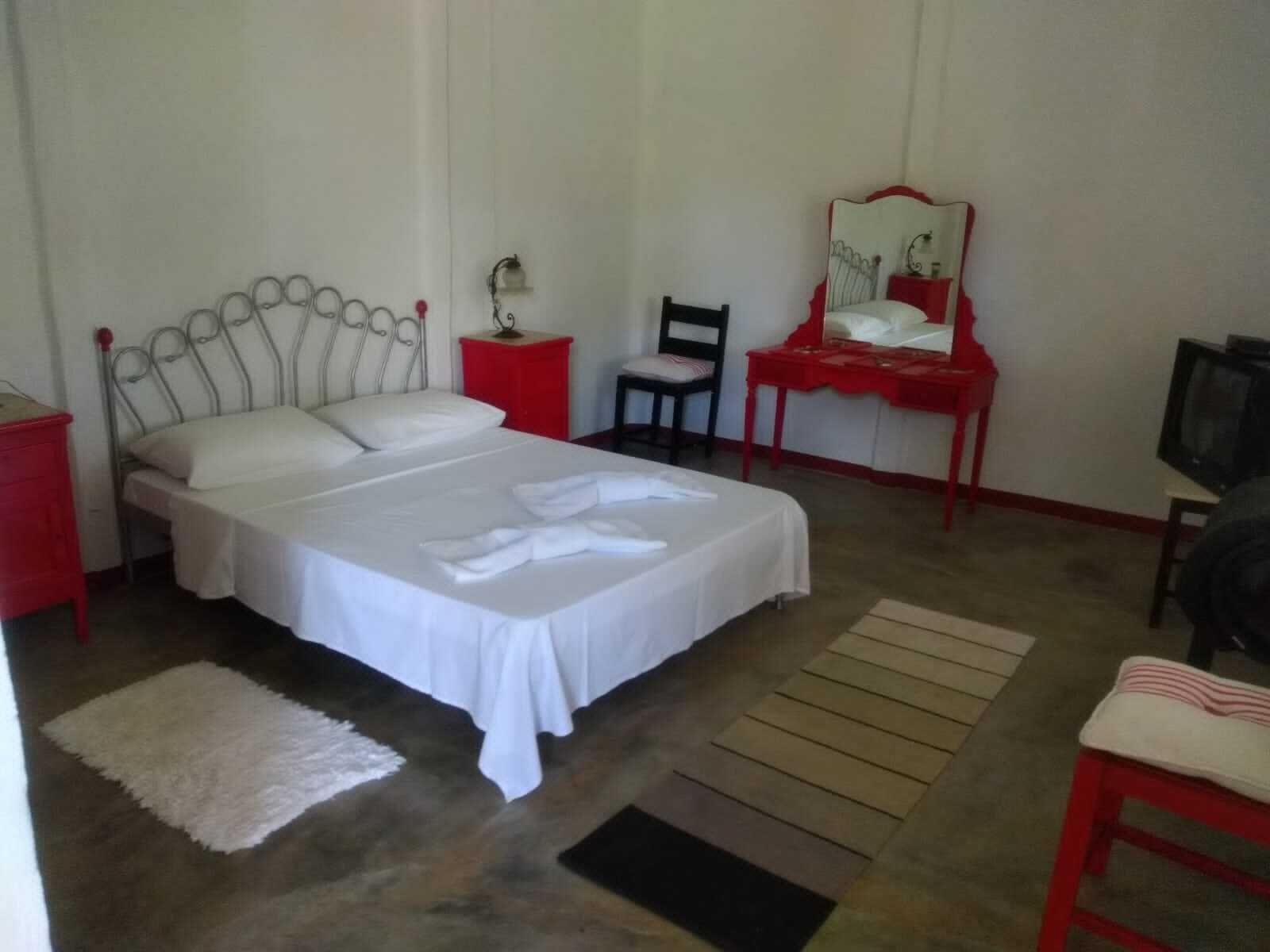 Une chambre avec un lit simple, une table de chevet rouge, une chaise, une petite table avec un miroir et un téléviseur.