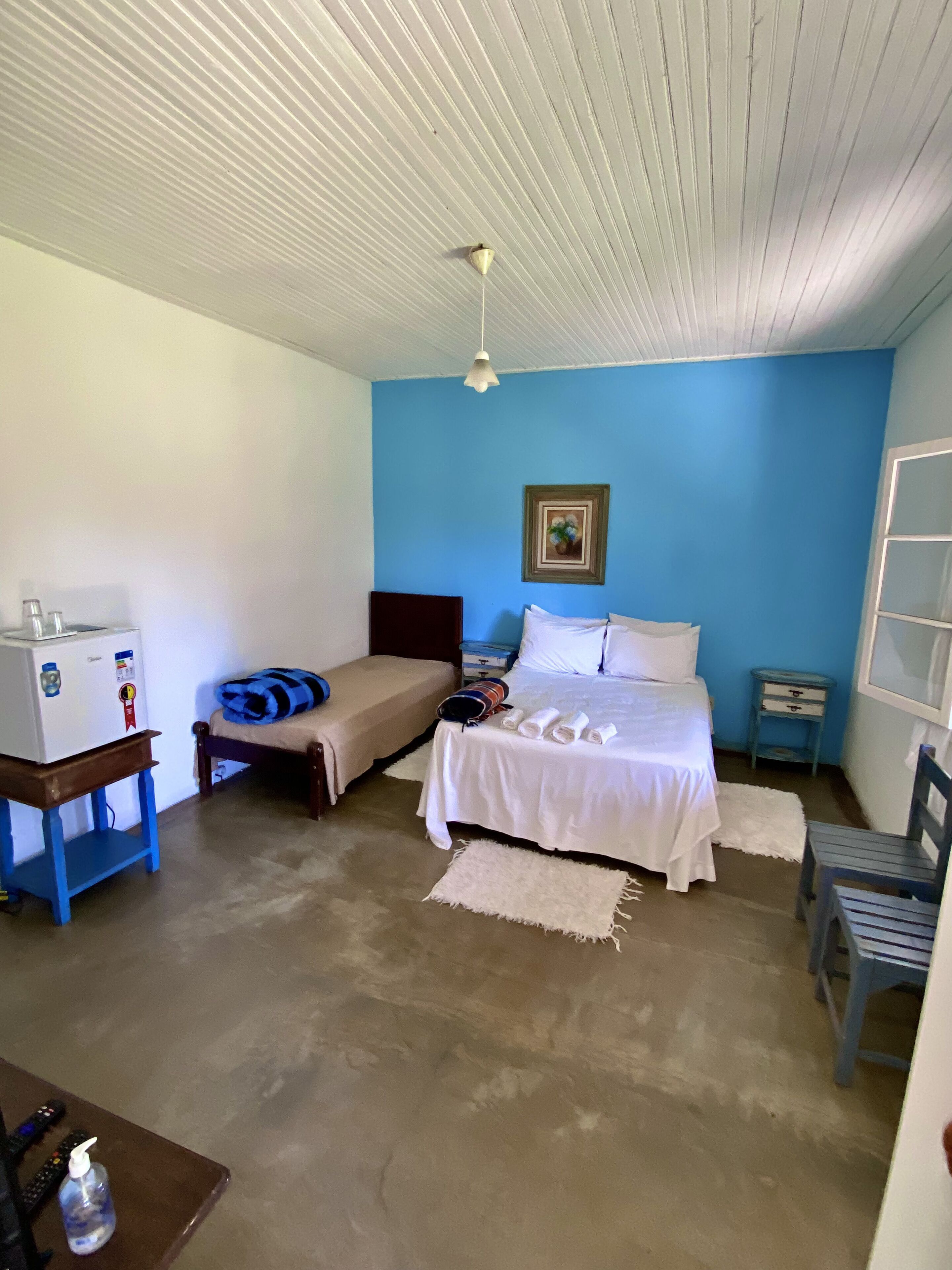 A compact room with a bed, a small table with a refrigerator, a chair, and a bedside table with a lamp.