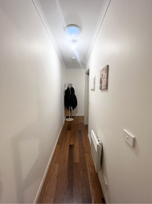 A narrow hallway with wooden flooring, white walls, a ceiling light, and a coat rack with jackets.