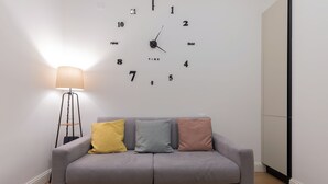 Een grijze bank met drie kussens in geel, blauw en roze, geplaatst tegen een muur met een grote klok die de tijd weergeeft.