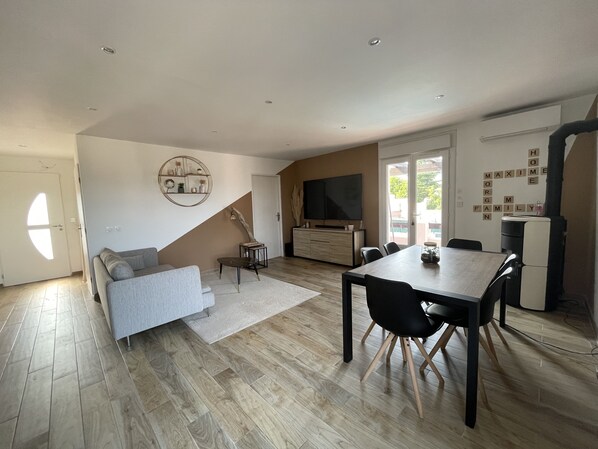 A modern living room with a grey sofa, a wooden dining table with black chairs, a TV, and a wall-mounted shelf.