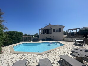 A swimming pool with stone tiles, a small building, and outdoor furniture.