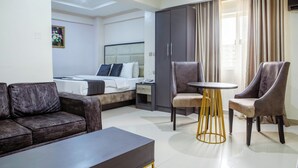 A hotel room with a bed, a sofa, two armchairs, a small table, a wardrobe, and a painting on the wall.