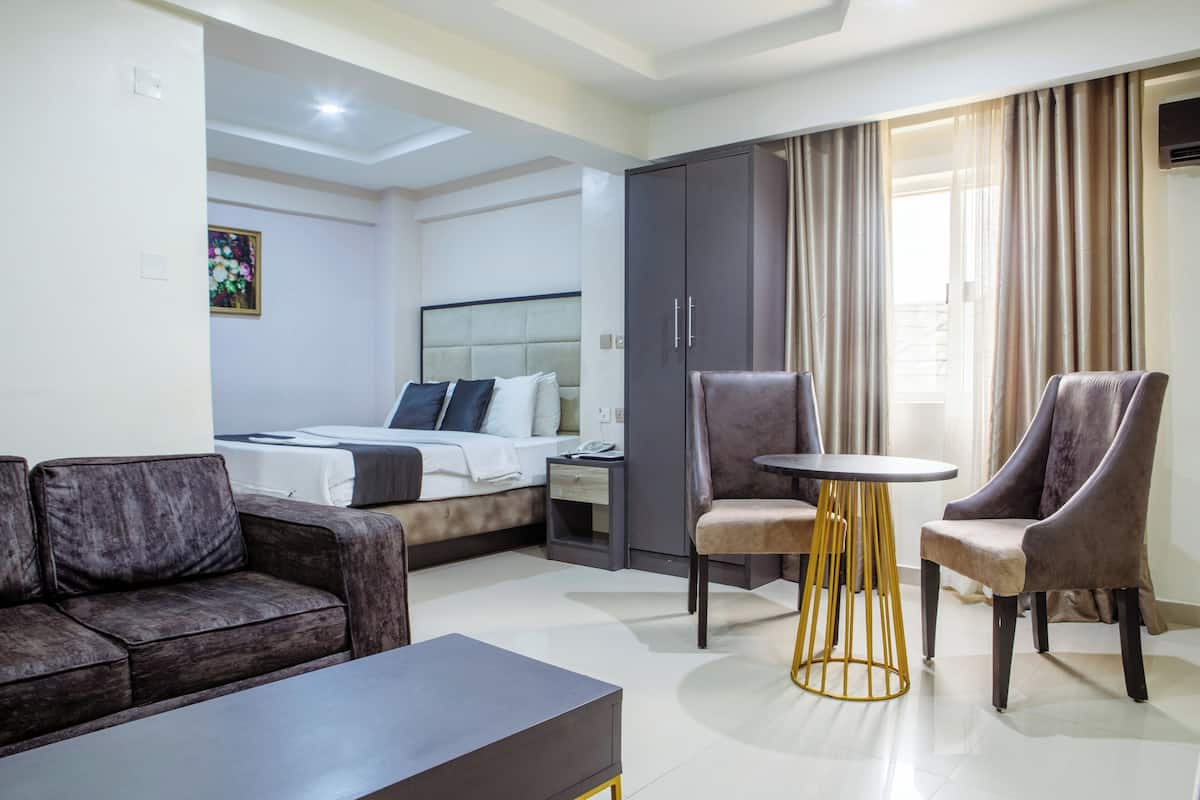 A hotel room with a bed, a sofa, two armchairs, a small table, a wardrobe, and a painting on the wall.