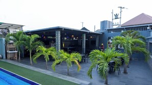 Un espace aménagé au bord de la piscine, avec un coin salon couvert, des palmiers et un bâtiment doté d’un toit en tuiles.