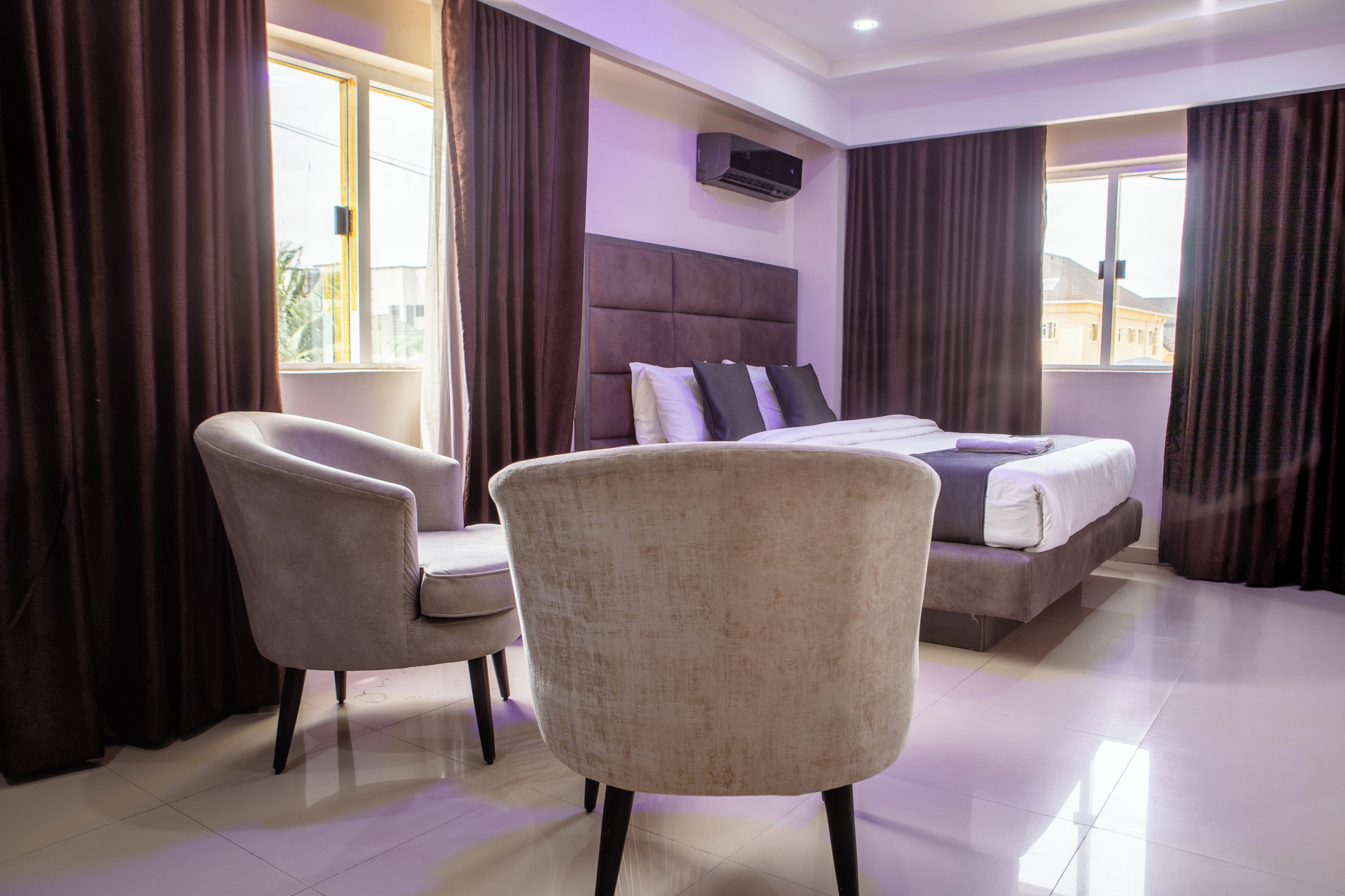 A hotel room with a bed, two armchairs, a window with curtains, and an air conditioning unit.