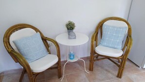 Due poltrone in vimini con cuscini bianchi e cuscini decorati con un motivo blu, un tavolino bianco con un vaso e una bottiglia, e un pavimento piastrellato.