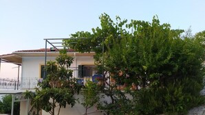 Una casa a due piani con un muro di sostegno in pietra, un balcone con ringhiere in metallo e un grande albero di fronte.