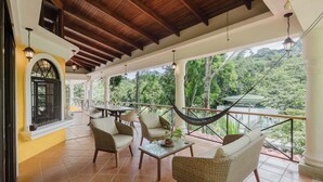A covered outdoor seating area with wicker furniture, a hammock, and a view of the surrounding greenery.