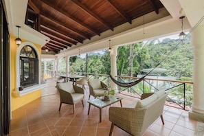 A covered outdoor seating area with wicker furniture, a hammock, and a view of the surrounding greenery.