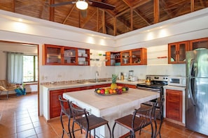 A spacious kitchen with wooden cabinets, a large island with chairs, a stainless steel refrigerator, and a ceiling fan.
