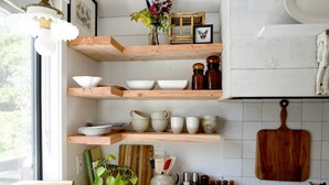 A kitchen with wooden shelves holding bowls and cups, a cutting board, and a vase with flowers.