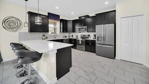 A modern kitchen with black cabinets, stainless steel appliances, a white island with bar stools, and a unique wall art piece. - Luxury 6 Bedroom 5 Bathroom Gated Resort Villa Close to Disney (Davenport)