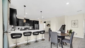 A modern kitchen with dark cabinets, a white countertop, and bar stools. There is a dining area with a round table and chairs. - Luxury 6 Bedroom 5 Bathroom Gated Resort Villa Close to Disney (Davenport)