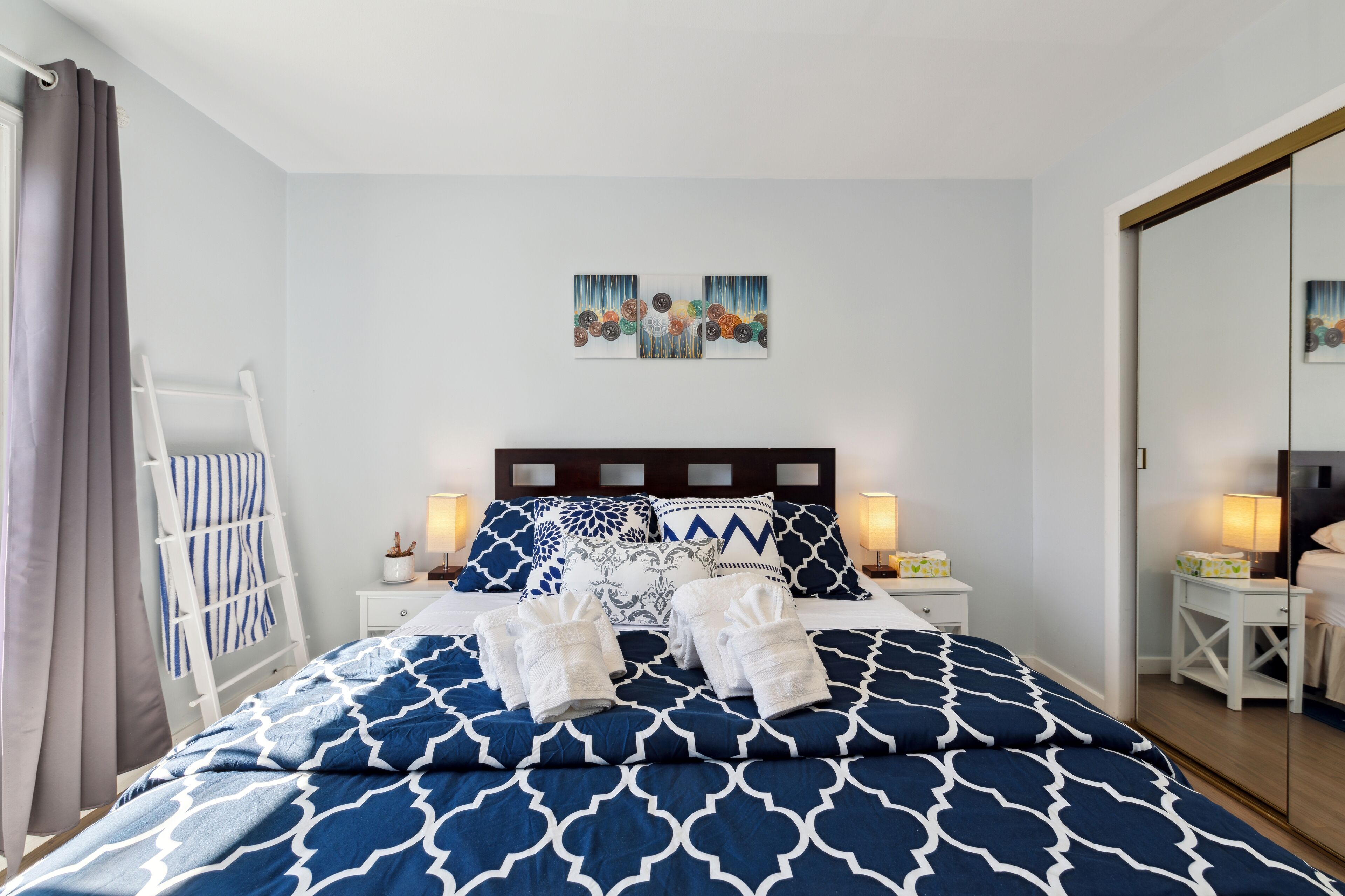 A neatly made bed with blue and white patterned bedding, two bedside lamps, a small bedside table with a lamp, and a wall-mounted artwork above the bed.