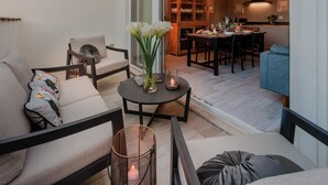 Ein modernes Wohnzimmer mit einer Couch, einem Sessel und einem kleinen Tisch mit Vase und Kerze. Im Hintergrund ist ein Essbereich mit Tisch und Stühlen zu sehen.