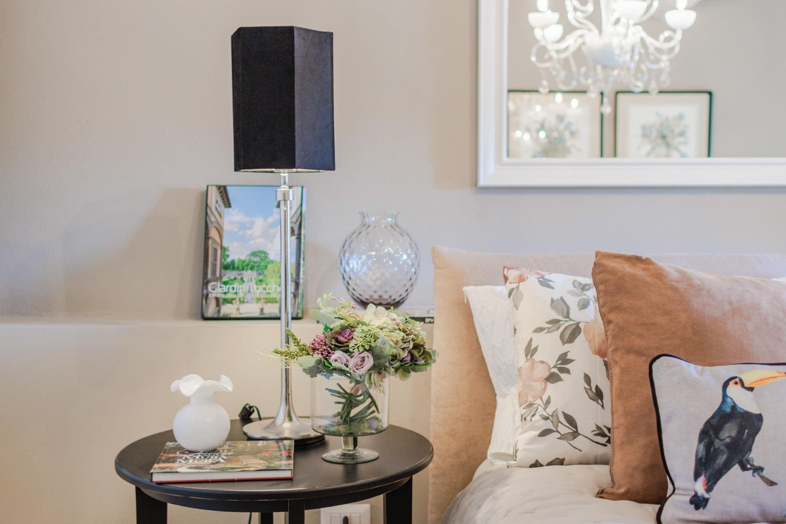A living room with a round table, a lamp, a vase with flowers, a book, and a couch with floral pillows.