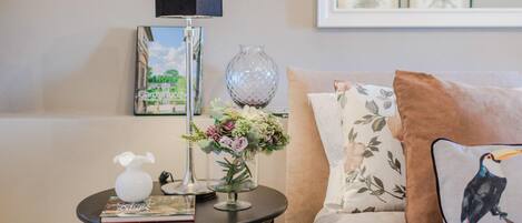 A living room with a round table, a lamp, a vase with flowers, a book, and a couch with floral pillows.