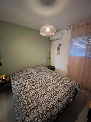 Un dormitorio con colcha estampada, lámpara colgante, aire acondicionado de pared y ventana con persianas.