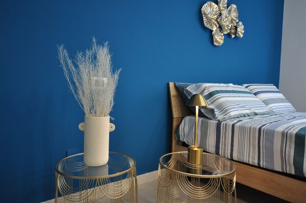 Un dormitorio con pared azul, cama con ropa de cama a rayas, mesita de noche con lámpara y decoración mural.