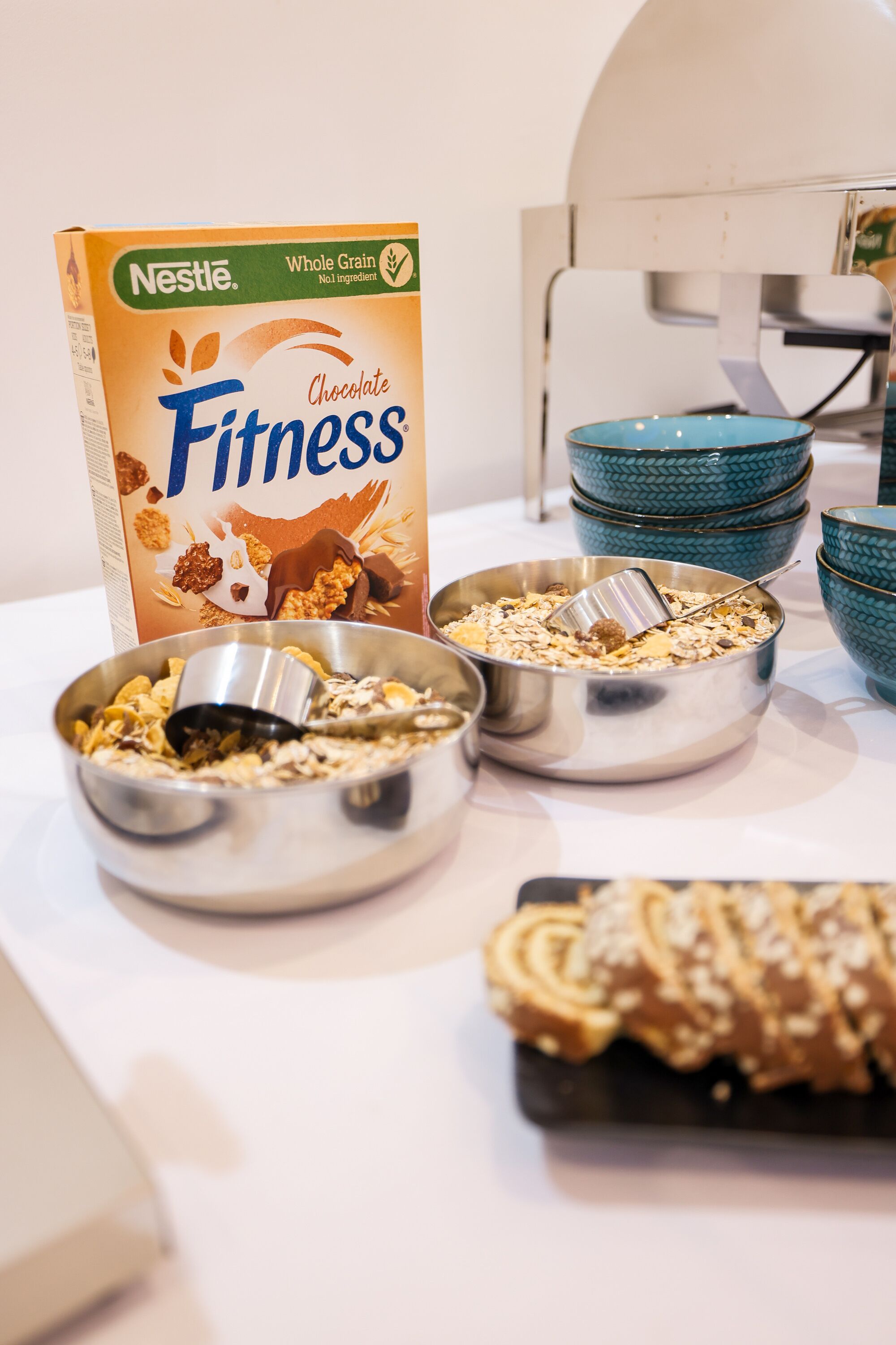 Een ontbijt opstelling met een doos Nestle Fitness chocoladevlokken, twee kommen met ontbijtgranen en melk, en een bord gebak.