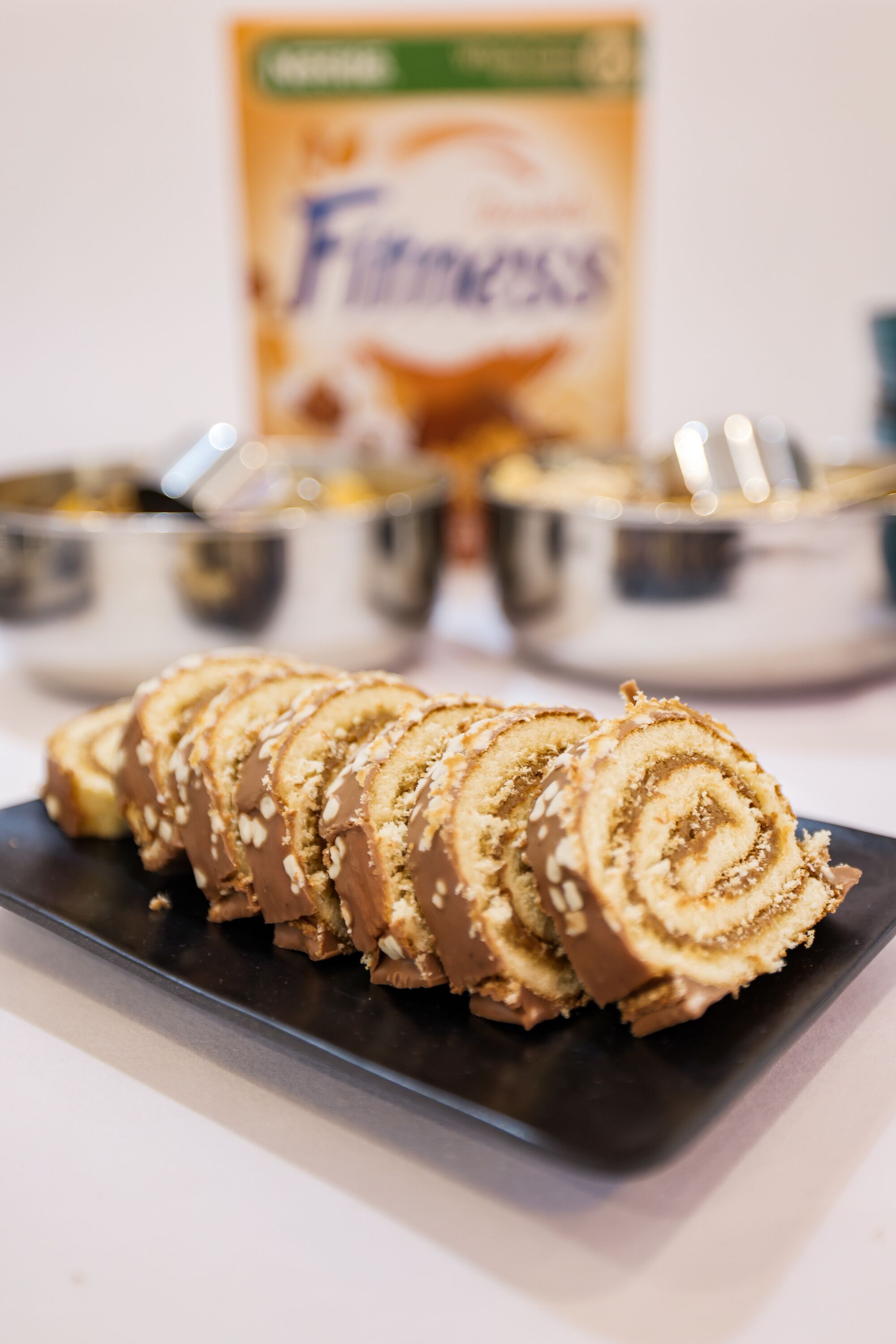 Ein Teller mit geschnittenen Brötchen mit Sesam, eine Schüssel Schokoladenaufstrich und eine Packung Fitness-Cereal im Hintergrund.