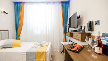A hotel room with a bed, a desk with a tray of fruit and wine glasses, a television, and a minibar.