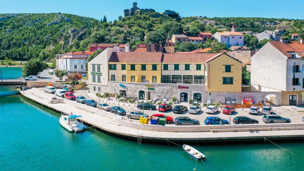 A coastal town with a marina, buildings with red-tiled roofs, and a hill with a castle-like structure in the background. - Hotel Kanjon Zrmanje (Obrovac)